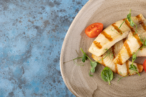 Grilled white Fish Fillet BBQ. tomato and microgreens on a plate on blue background. Copy space. Top view