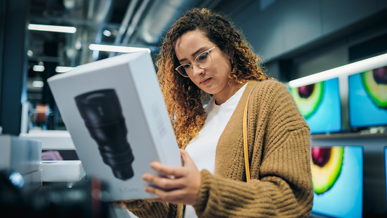 Beautiful Latin Female Looking at a Box with a Modern Photo Lens, Reading Model's Specifications and Features. Young Client Shopping for a Camera Zoom Lens in a Home Electronics Store