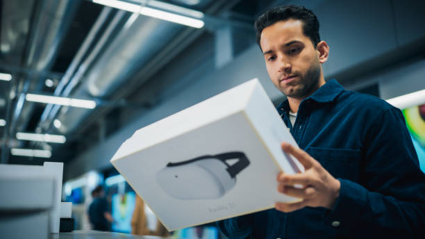 handsome young man shopping for a virtual reality computer in a home electronics store. multiethnic customer comparing different headset technology features, sale price and vr quality of the device - department store clothing showroom people ストックフォ�トと画像