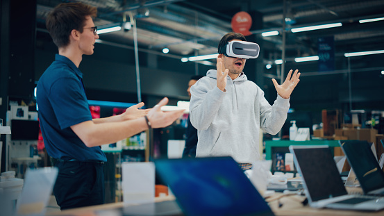 Young Man Having Conversation with a Salesperson in an Electronics Store while Trying Out a Virtual Reality Device. Customer Wishes to Buy a VR Set. Shopper Explores Latest Options with Expert Advice