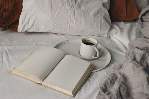 Cup of coffee, open books on white bed sheet. Breakfast in bed concept. Blurred muslin, linen pillows, blanket. Moody cozy Scandinavian bedroom. Top view, no people, lifestyle banner.