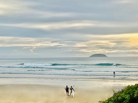 Horizontal seascape of surfers talking on sand with breaking waves under a cloudy overcast sky with distant island and foreground coastal tree top at Emerald Beach NSW Australia