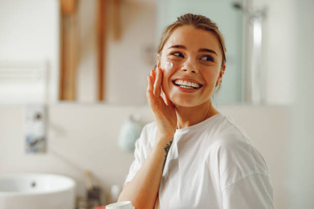 Young beautiful woman taking care of skin by applying moisturizer cream in bathroom Happy woman applying hydrating moisturizer on her face sitting in bathroom. Home beauty routine suntan lotion stock pictures, royalty-free photos & images