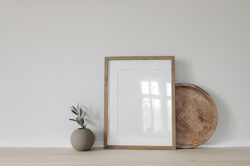 Blank vertical picture frame mockup, poster display. Ball vase with olive tree branch. Round wooden tray on table, desk. Minimal rustic home. Scandinavian interior, white wall background.