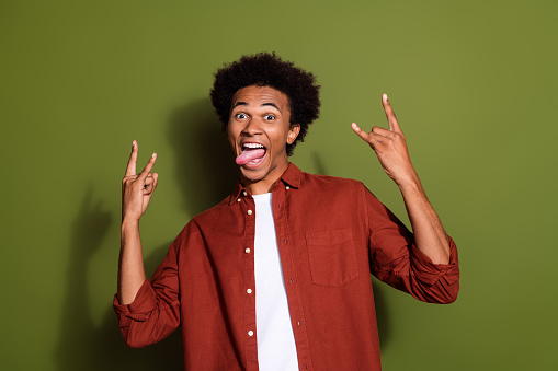 Photo portrait of handsome young guy show rock gesture tongue out dressed stylish brown outfit isolated on khaki color background.