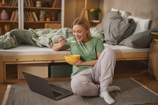 Happy young woman using laptop while having breakfast in bedroom