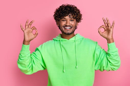 Photo of friendly cheerful young guy model in green sweatshirt showing okey sign double like symbol isolated on pink color background.