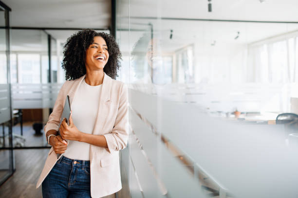 A young professional woman stands confidently in a modern office Brazilian businesswoman with an infectious smile standing in a modern office corridor and holding her laptop. Cheerful female employee satisfied with her occupation, feels happy and successful professional woman smiling stock pictures, royalty-free photos & images