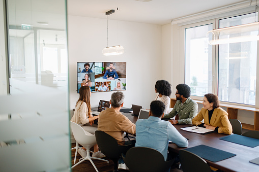 In a well-lit meeting room, a team actively engages with remote colleagues via a large screen, illustrating the modern hybrid work model where technology bridges the gap between in-office and remote team members.