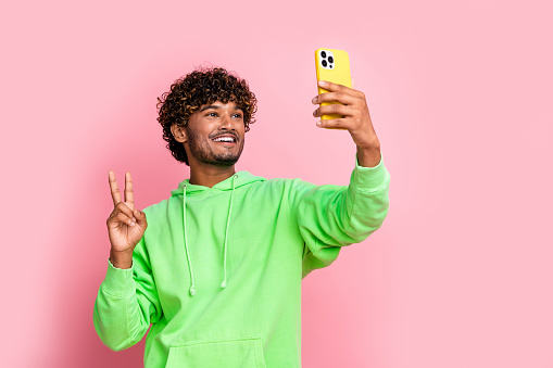 Self portrait of young blogging arabian guy holding smartphone shows v sign into cadre wear hoodie isolated on pink color background.