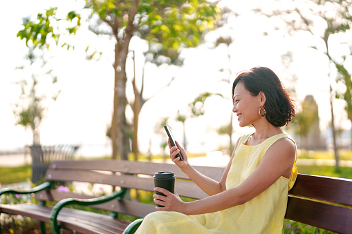 Portrait of a young Asian woman using a smartphone while enjoying a cup of coffee in the park, savoring a beautiful day outdoors and breathing in the fresh air.