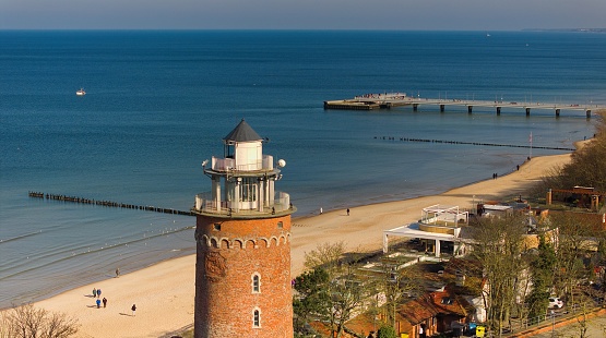 February in Koobrzeg, Poland: A serene scene featuring a red brick lighthouse, people enjoying a sunny day on the sandy beach, and a tranquil sea without any waves.