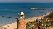 A sunny day in February at Koobrzeg port, Poland. The image captures a red brick lighthouse, tourists strolling on the sandy beach, and a distant pier, with calm sea waters.