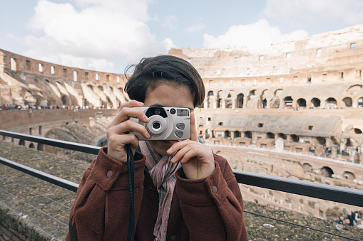 A girl is taking a picture with camera film of the Roman Colosseum,  the girl is wearing a brown jacket