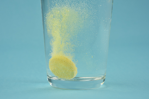 Close up photograph of orange colour water soluble vitamin C dissolving in glass of water with bubbles on blue colour background