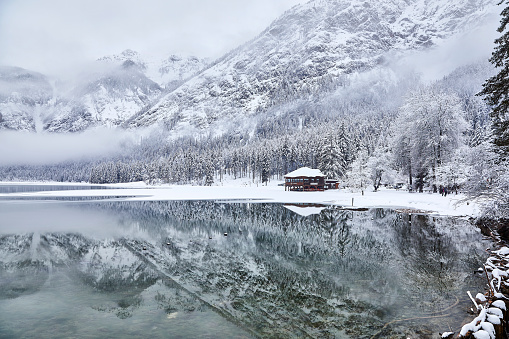 Lago Di Dobbiaco in winter with a perfect reflection, Dolomites, Italy everything is covered with snow and the mist is hanging above the lake. There is a cabin standing at the lake.