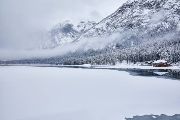 Lago Di Dobbiaco in winter, Dolomites, Italy everything is covered with snow and the mist is hanging above the lake. There is a cabin standing at the lake.