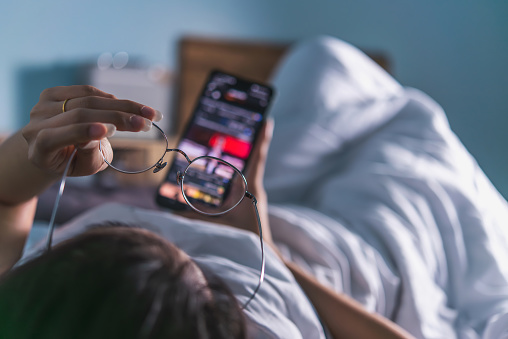 An over-the-shoulder view of a person in bed checking a smartphone with glasses in hand, signifying a modern morning routine.