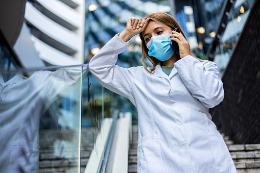 Medical doctor woman talking on phone outside near hospital. Modern medical doctor woman in uniform and medical mask speaking on a smartphone outside near hospital.