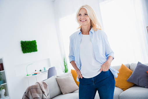 Photo of smiling cheery successful business woman blonde mature aged wearing blue shirt looking interested standing home in living room.