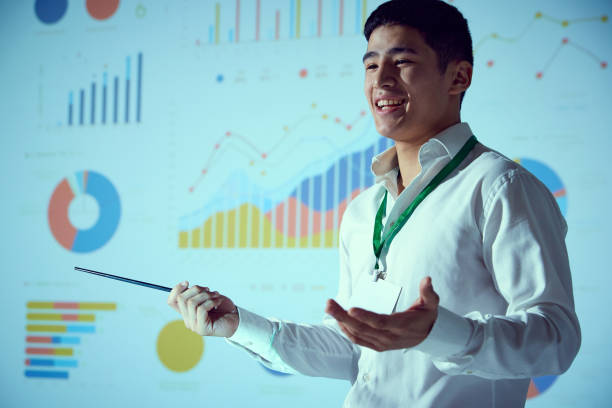istock Asian businessman, speaker delivering presentation to attentive crowd with projection of colorful statistical graphs behind. 2065766641