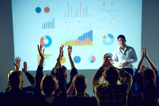 corporate training session surrounded by data visualizations where man ending speech and audience clapping raising hands. - training business seminar clapping imagens e fotografias de stock