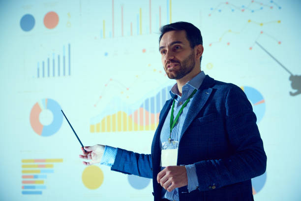 istock Business conference scene featuring businessman, presenter addressing audience with visual aid of pie charts and bar graphs. 2065766655