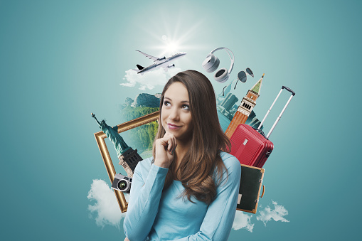 Happy woman dreaming of traveling, she is surrounded by travel items and thinking with hand on chin