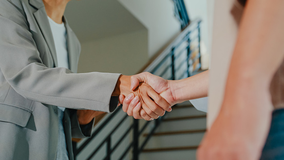 Close-up of Asian female real estate agent handshaking after complete agreement with tenants after showing new houses. Landlord selling real estate to happy couple customer for investment concept.