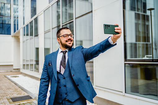 Young smiling businessman taking selfies in front of an office building