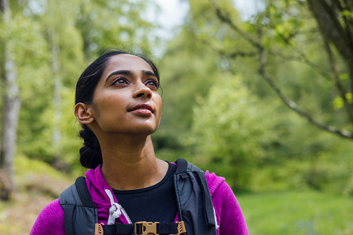 Close up front view of a young woman stood in athletic leisure. She is grounding and enjoying the fresh air and green surroundings and is feeling calm and serene. She is looking up into the distance.