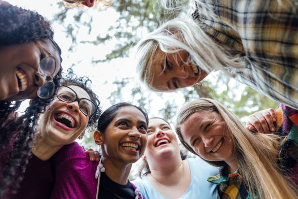 Low angle view of a medium group of friends standing together in a huddle with their arms around each other in a wooded area in Northumberland, North East England. They are talking before going on a hike in the countryside, wearing casual clothing.