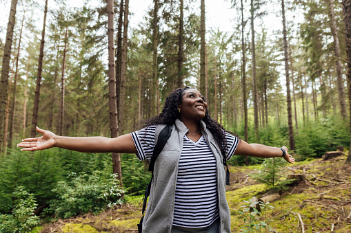 Waist up front view of a young woman stood in athletic leisure. She is grounding and enjoying the fresh air and green surroundings and is feeling calm and serene. She has her arms outstretched and doing a breathing exercise.