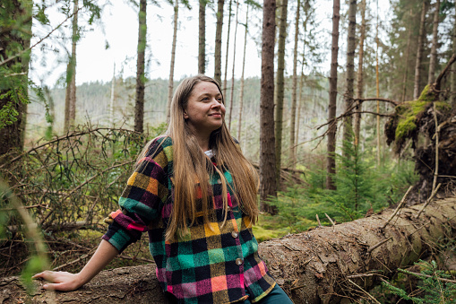 A waist up view of a young woman stood grounding and enjoying the fresh air and green surroundings and is feeling calm and serene. She has her hands resting on a fallen tree and is doing a breathing exercise.