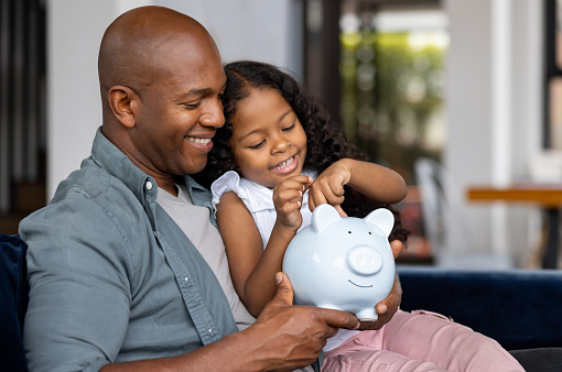 Loving African American father and daughter saving money in a piggybank - financial literacy concepts