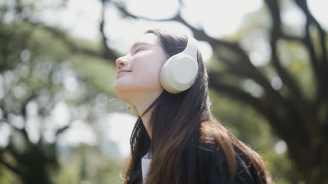 Side view of Cheerful young woman listening to music with big trees in the background.