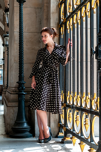 Full length shot of a young brunette in a dotted polka dress on high heels leaning against classic iron fence in Paris. Full body shot.
