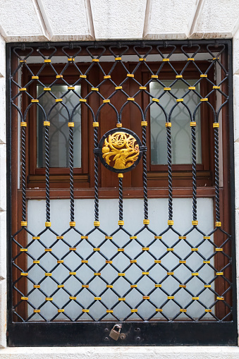 Image of a window with ornate Venetian style security grill. The grill has a gold central boss depicting a tree. The top an bottom halves of the grill have geometric patters and are separated by twisted metal rods. The grill is padlocked to the frame.