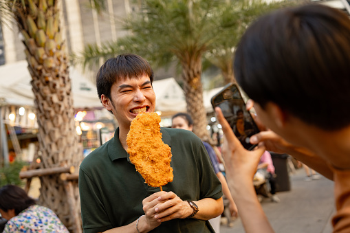 Joyful Asian couple vlogging with a fried chicken at the street food market on the weekend.