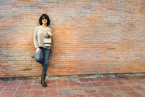 Smiling woman leaning on a brick wall with casual clothes and handbag