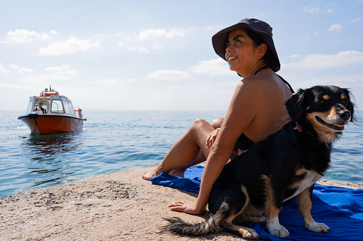 Latin woman wearing a hat and bikini and enjoying a beautiful sunny morning on a jetty with her pet on the Greek island of Crete