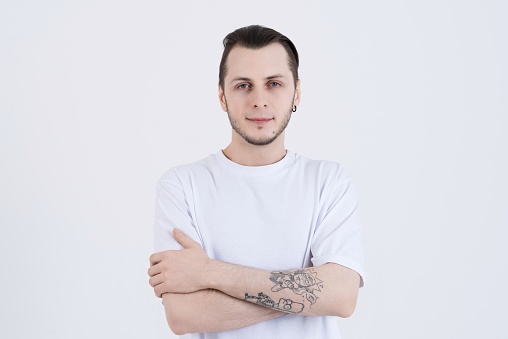 Portrait of an attractive Caucasian young man with tattoos in casual clothes with crossed arms isolated on white studio background. Confidently looking at the camera