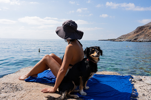 Unknown woman sitting sunbathing in a bikini and wearing a hat next to her pet by the sea on a beautiful summer day on the Greek island of Crete