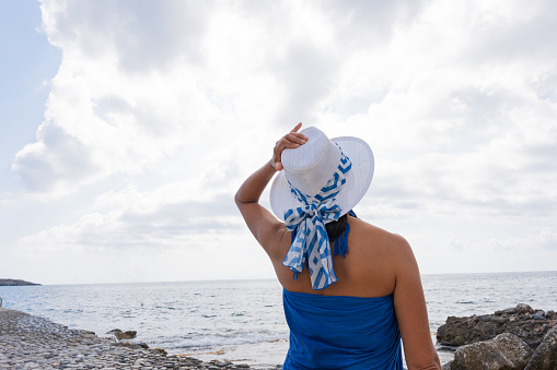 Woman from behind in a blue sarong holding her hat and enjoying the views of the sea from a Greek tavern in the south tavern in the south of the island of Crete