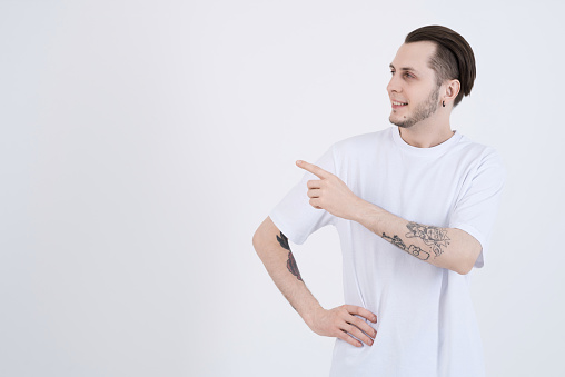 Attractive caucasian young man with tattoos in casual clothes smiling and pointing finger at blank copy space for advertising isolated on white studio background.