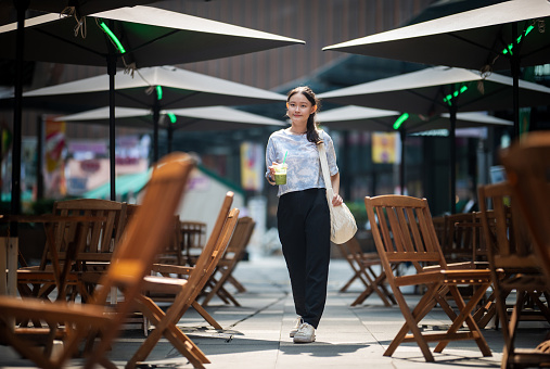 A young smiling Asian woman strolls past a roadside café, enjoying drinks during her travels.