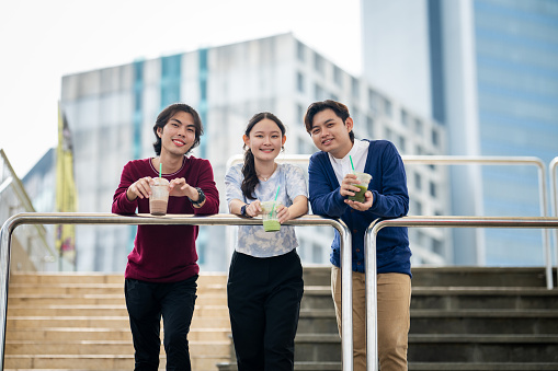 A group of cheerful young Asian friends stand on the stairs, relishing drinks, laughter, and outdoor fun while on their travels. Looking at camera.
