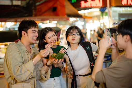 Asian LGBTQ couple and friend exploring Bangkok's night market, indulging in popular street food delights.