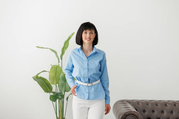 Women equality day. Smiling successful businesswoman in elegant blue shirt and white pants looks at camera at modern comfortable light office with green plant and sofa. Work life balance concept día de la igualdad de la mujer. sonriente mujer de negocios exitosa con elegante camisa azul y pantalones blancos mira a la cámara en una oficina moderna y cómoda con luz con planta verde y sofá. concepto de equilibrio entre la vida laboral y personal.