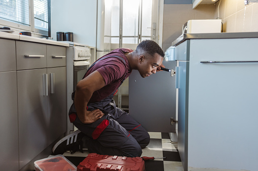 Photo of African American handyman in work clothes fixing leak in the kitchen sink